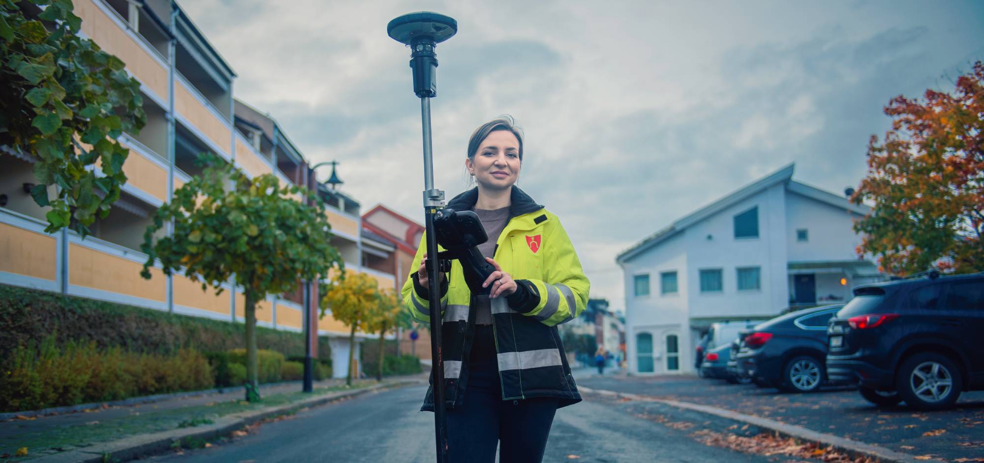 En landmåler i gul refleksvest holder et GPS-målepunkt på stativ i en rolig bygate om høsten.