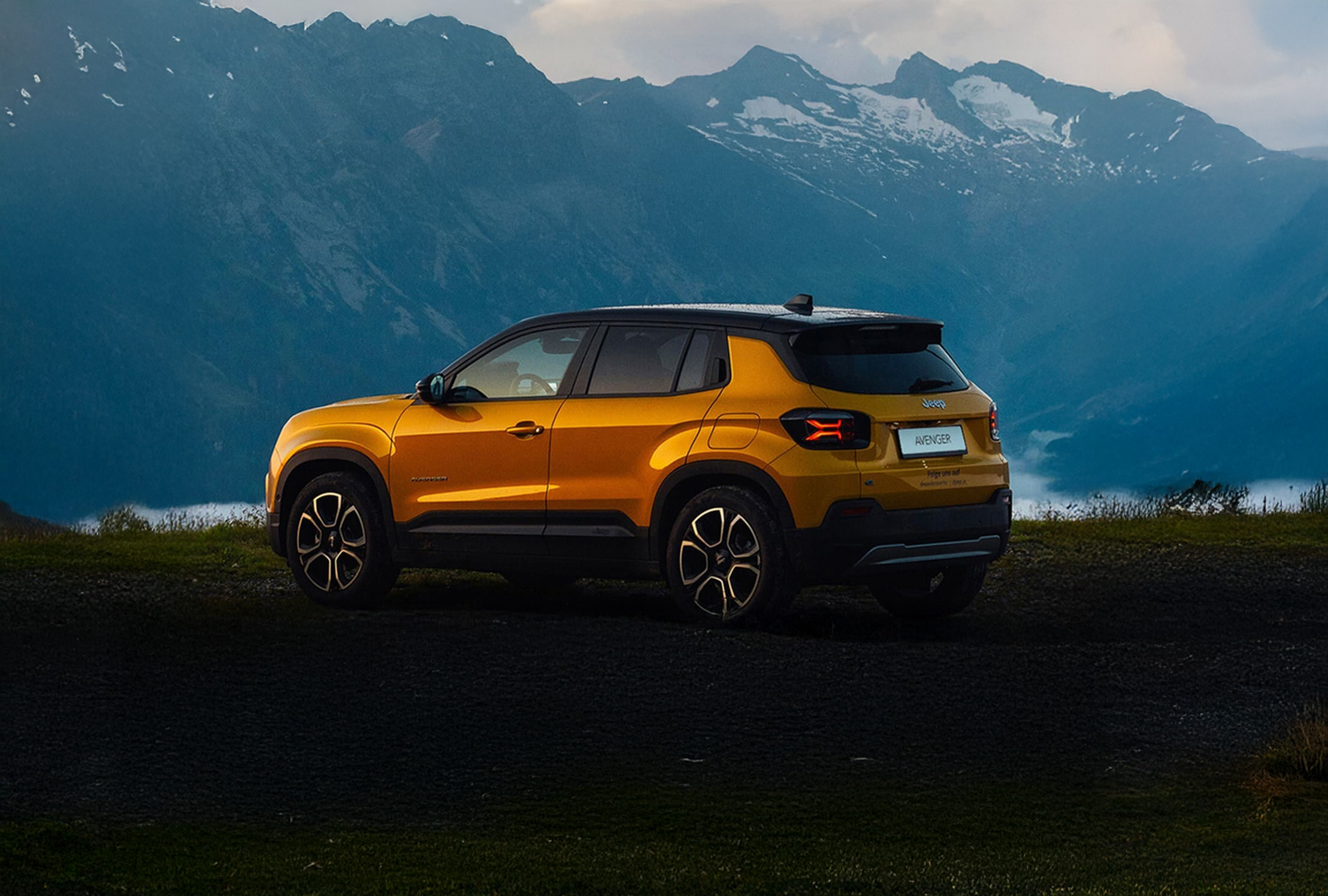 En gul Jeep Avenger parkert i et dramatisk fjellandskap med mørk bakke og blå fjell i bakgrunnen.